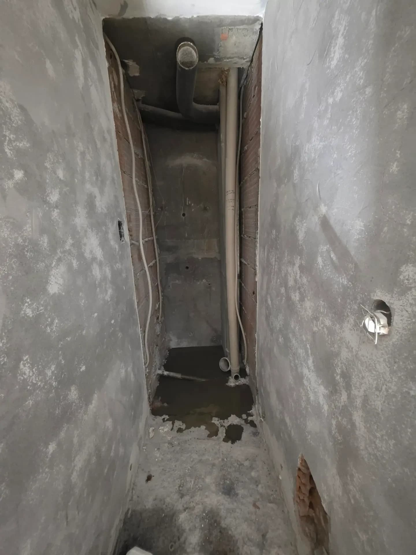 WC étroit en rénovation à Méribel, murs enduits gris, tuyaux et gaines visibles au sol, vue vers l’extérieur.
