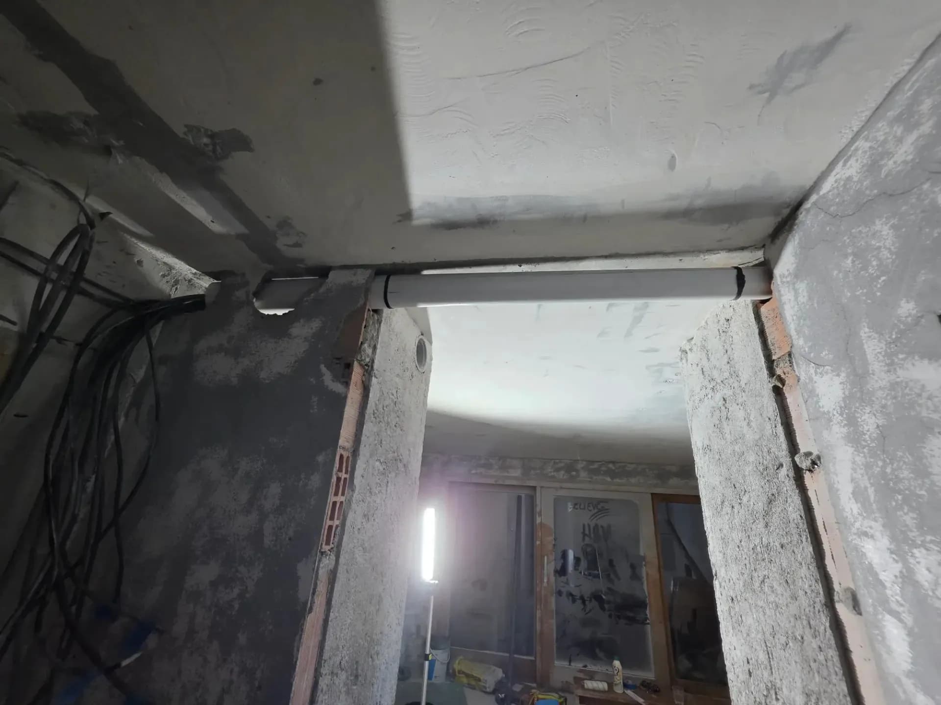 Plafond et murs en construction, tuyau blanc traversant, câbles et gaines visibles dans un couloir.