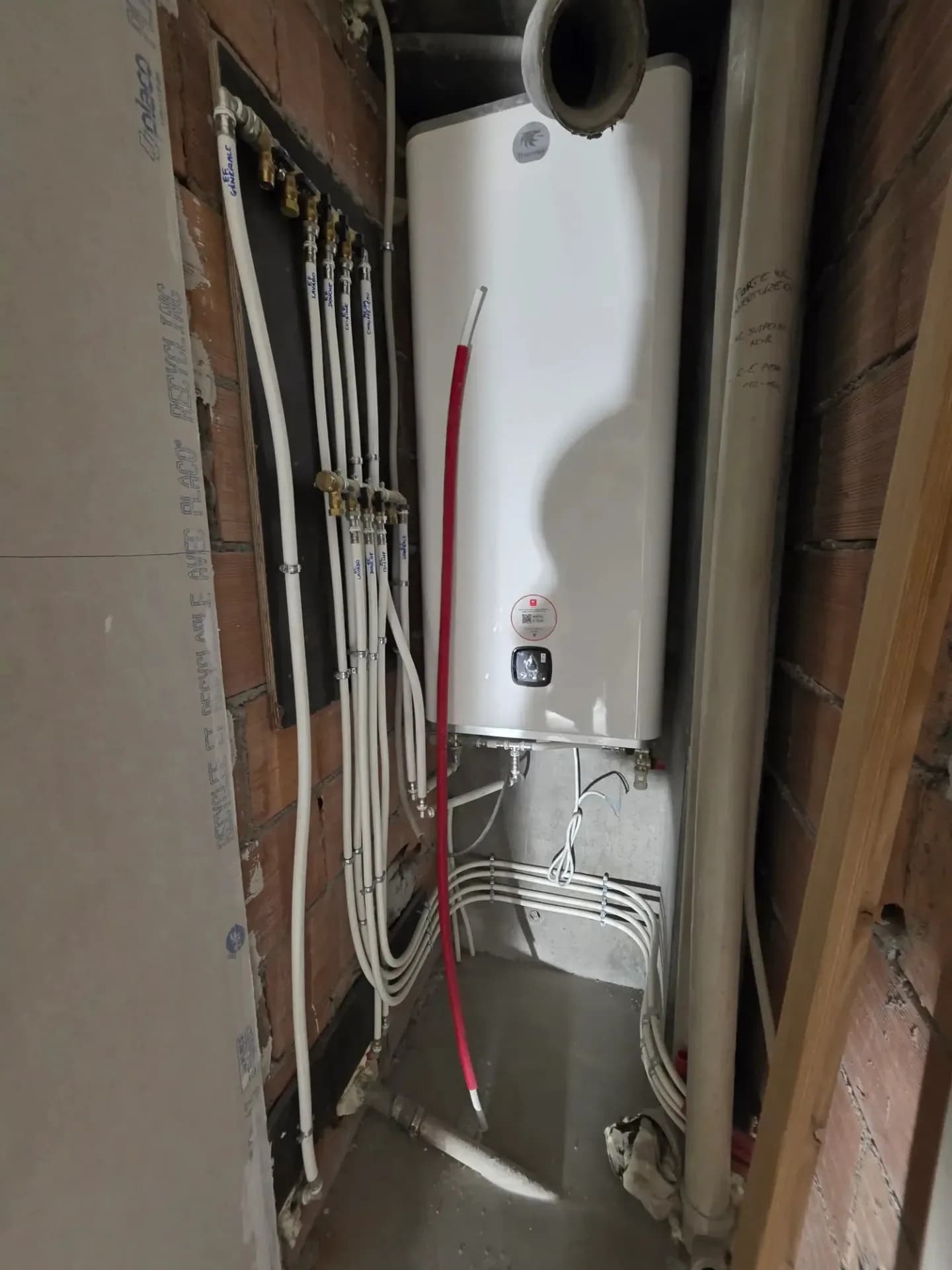 Chauffe-eau blanc installé dans un local, avec tuyaux blancs et rouges, câbles et raccords visibles.