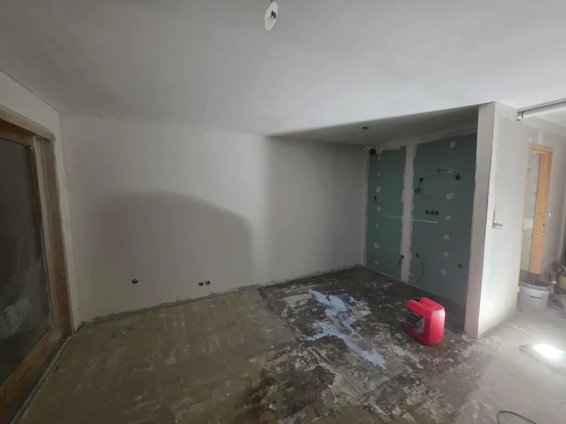 Salon en rénovation, murs blancs enduits, sol poussiéreux, pièce ouverte sur le couloir.
