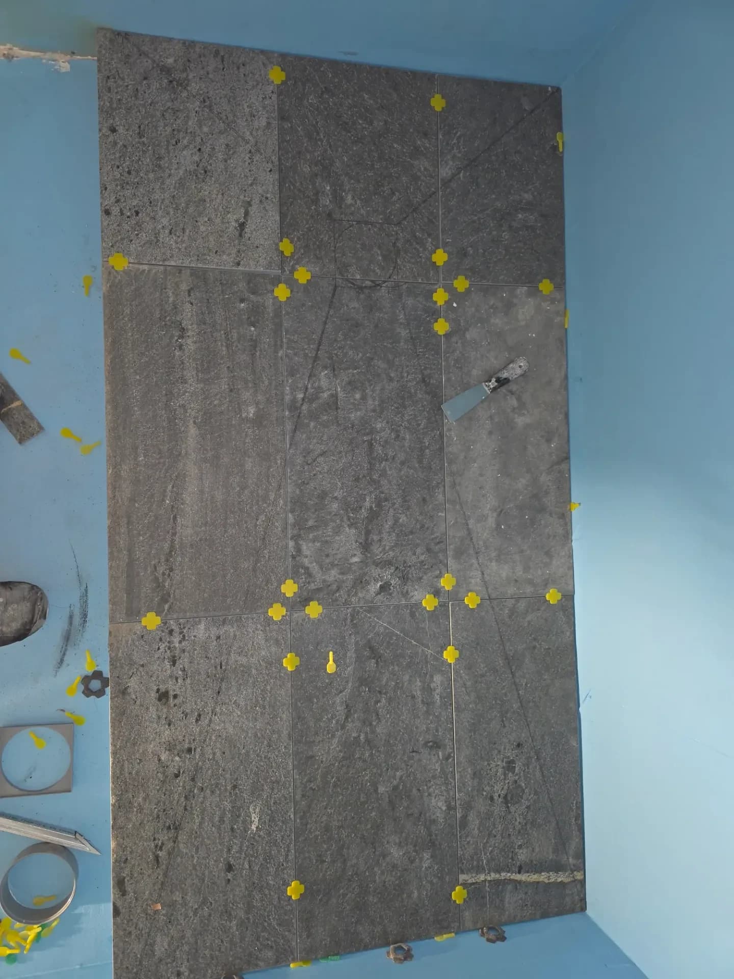 Carrelage gris effet ardoise texturé, avec croisillons jaunes et outils de pose sur le fond bleu de l'étanchéité.
