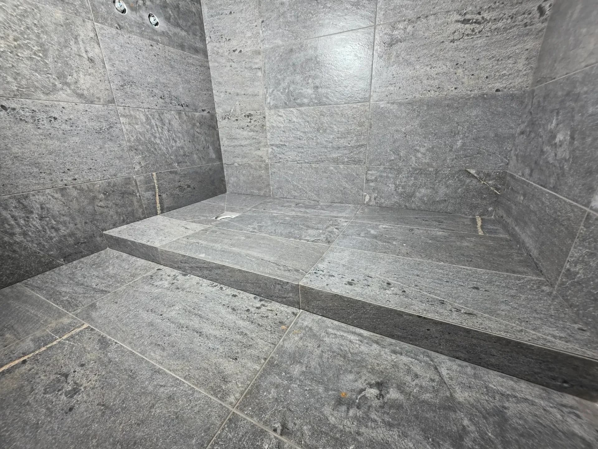 Douche à l’italienne carrelée gris, receveur rectangulaire avec bonde, murs en carreaux et joints visibles.