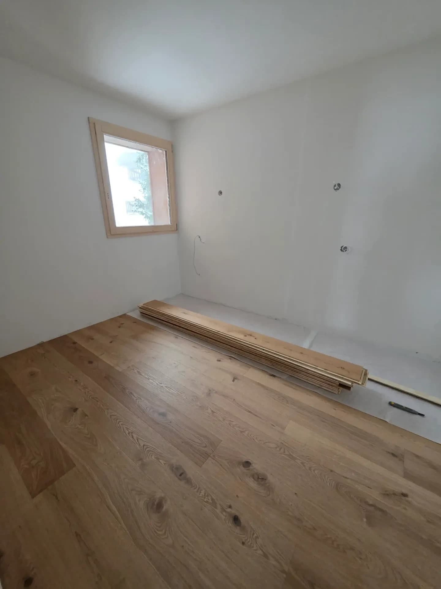 Pièce vide aux murs blancs, fenêtre en bois, câbles électriques et lames de parquet empilées au sol.