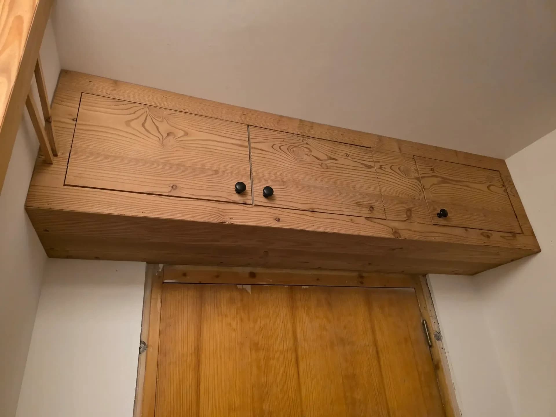Meuble en bois mélèze avec portes ouvrantes et poignées noires, installé au-dessus d’une porte d'entrée.